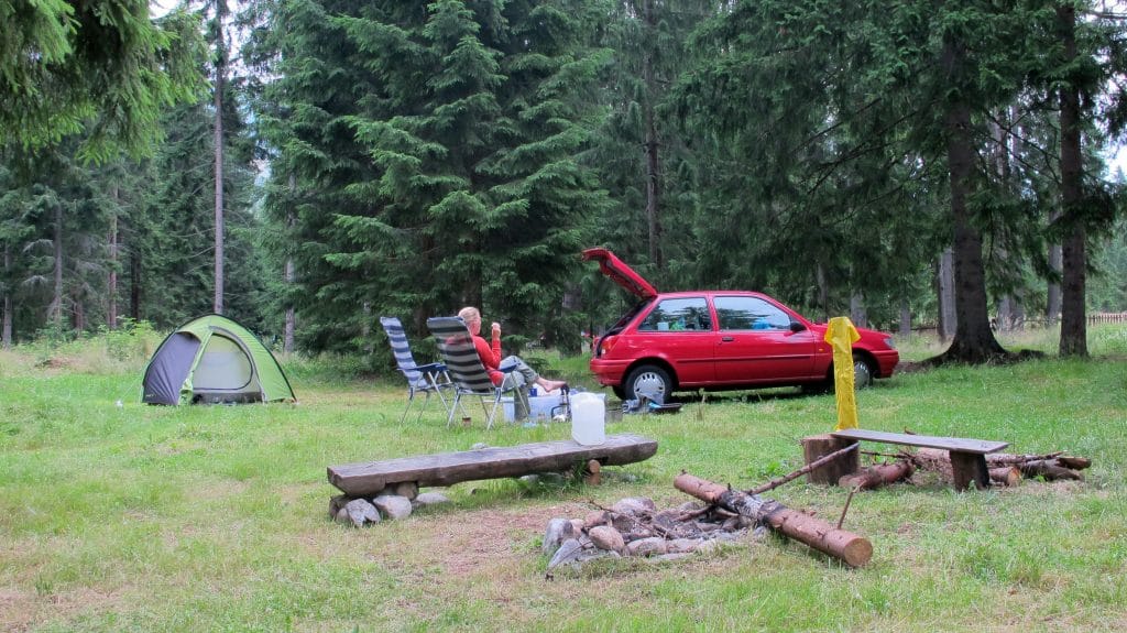 Persoon ontspant in campingstoel naast groene tent en rode auto in het dennenbos op camping Rackova Dolina bij Jamník, Slowakije