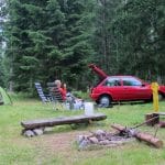 Persoon ontspant in campingstoel naast groene tent en rode auto in het dennenbos op camping Rackova Dolina bij Jamník, Slowakije