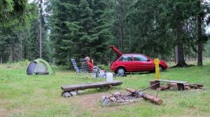 Persoon ontspant in campingstoel naast groene tent en rode auto in het dennenbos op camping Rackova Dolina bij Jamník, Slowakije