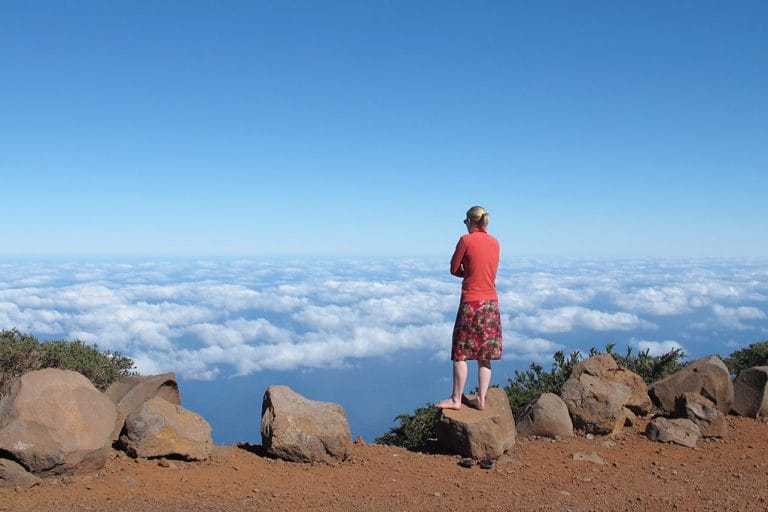 Vrouw staat op een rotsachtige bergtop boven een wolkendek op Roque de los Muchachos, La Palma, met een blauwe lucht