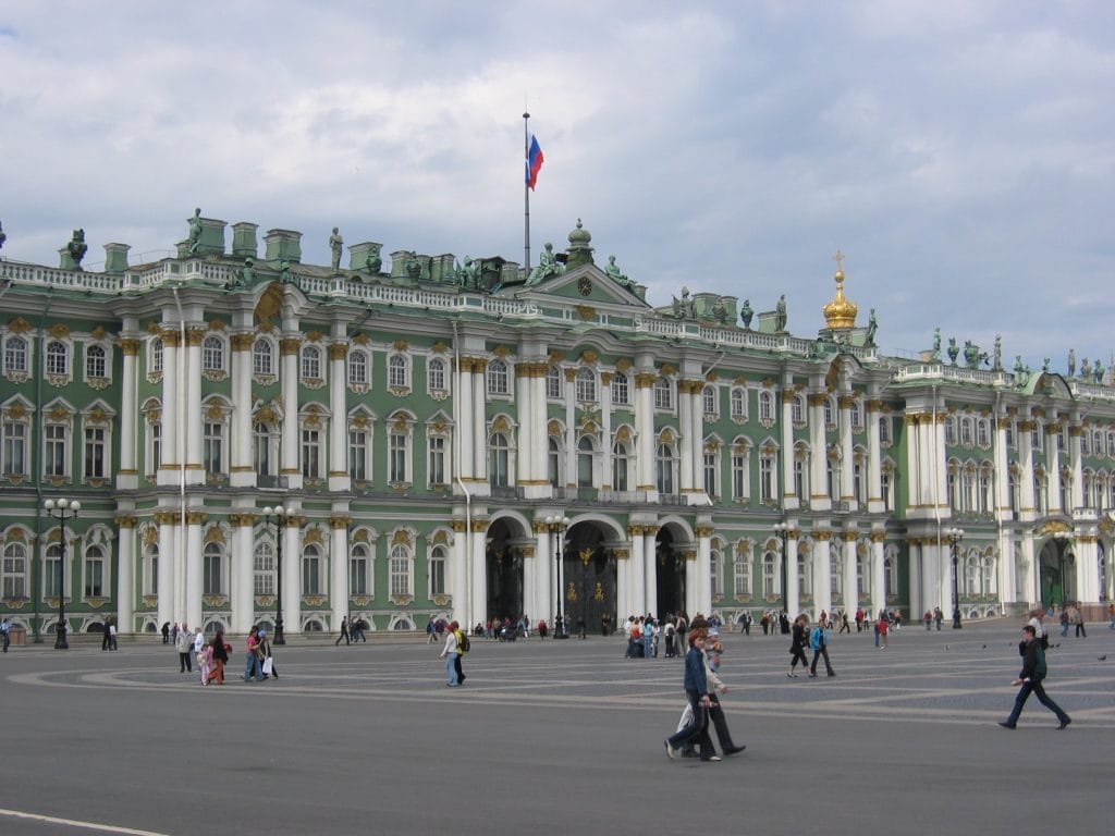 Hoofdingang van de Hermitage in St. Petersburg met de Russische vlag op het dak en gouden koepels van een orthodoxe kerk op de achtergrond