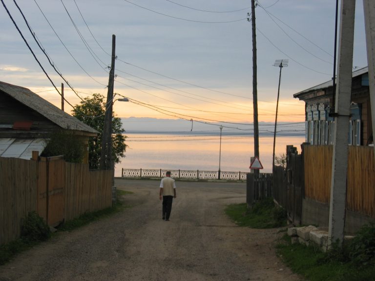 Smal straatje in Listvyanka met houten hekken en elektriciteitspalen en zicht op het Baikalmeer bij zonsondergang