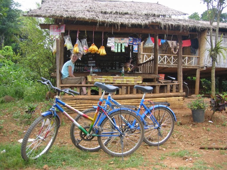 Fietser houdt pauze bij een kleine Laotiaanse dorpswinkel op palen onder een rieten dak, met hangende zakjes snacks en fietsen op de voorgrond