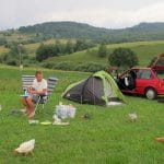 Man kookt op campingstoel naast groene tent en rode auto met kippen op de voorgrond op camping Sedliacky Dvor in Rohozna, Slowakije