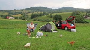 Man kookt op campingstoel naast groene tent en rode auto met kippen op de voorgrond op camping Sedliacky Dvor in Rohozna, Slowakije