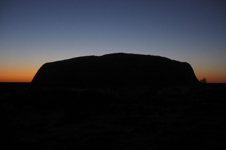 Uluru als donker silhouet tegen een gradientlucht van oranje naar diepblauw bij zonsopkomst in het Northern Territory van Australië
