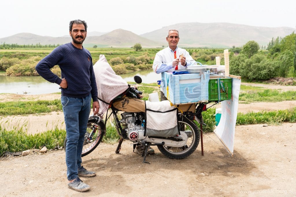 Twee mannen bij een mobiele straatverkoopkar langs een rivier in het Iraanse platteland, met groene heuvels en bergen op de achtergrond
