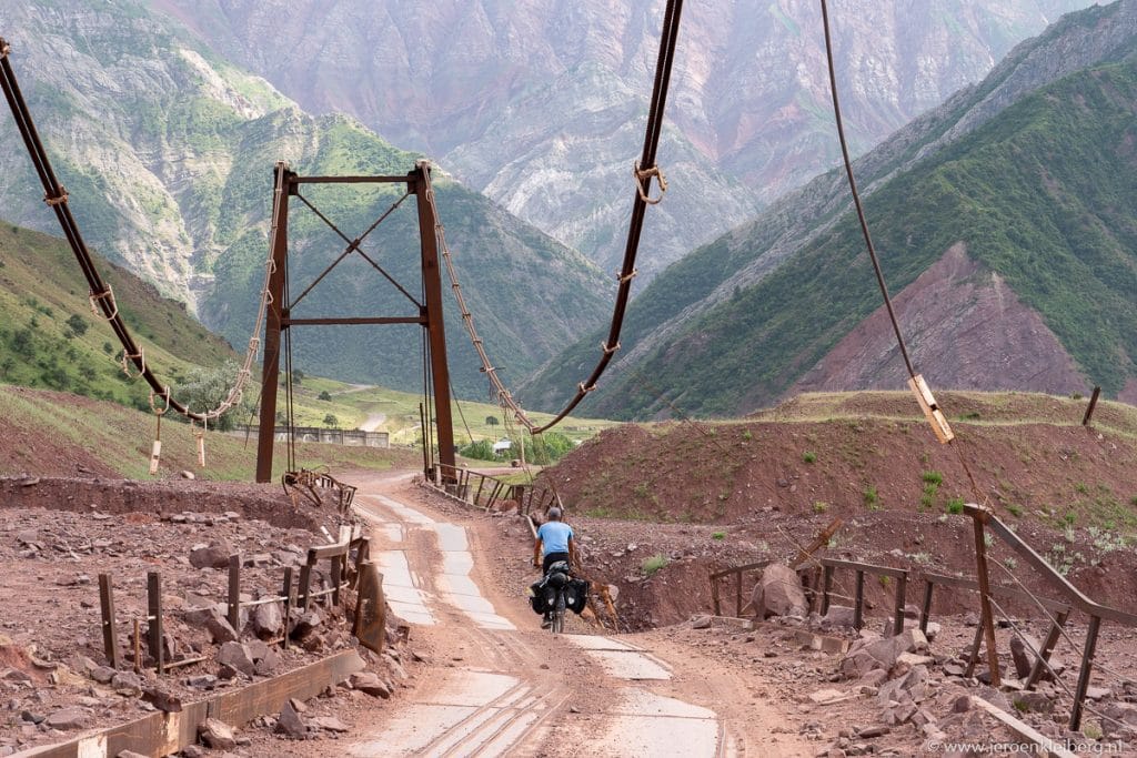 Fietser met fiets op een roestige wankele hangbrug over een diepe rotskloof in Tadzjikistan met dramatische bergwanden op de achtergrond