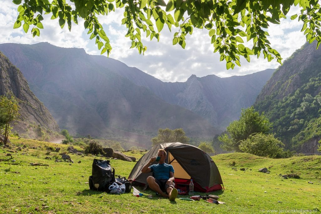 Persoon zit voor een tent op een groene kampeerplaats in een vallei in Tadzjikistan, omgeven door hoge bergwanden en weelderige bomen