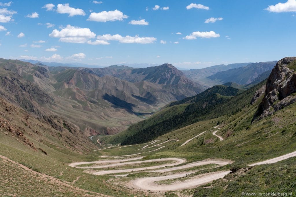 Kronkelende onverharde weg met meerdere bochten door een groene berghoogvlakte in Kirgizistan, met wijds uitzicht op omliggende bergtoppen