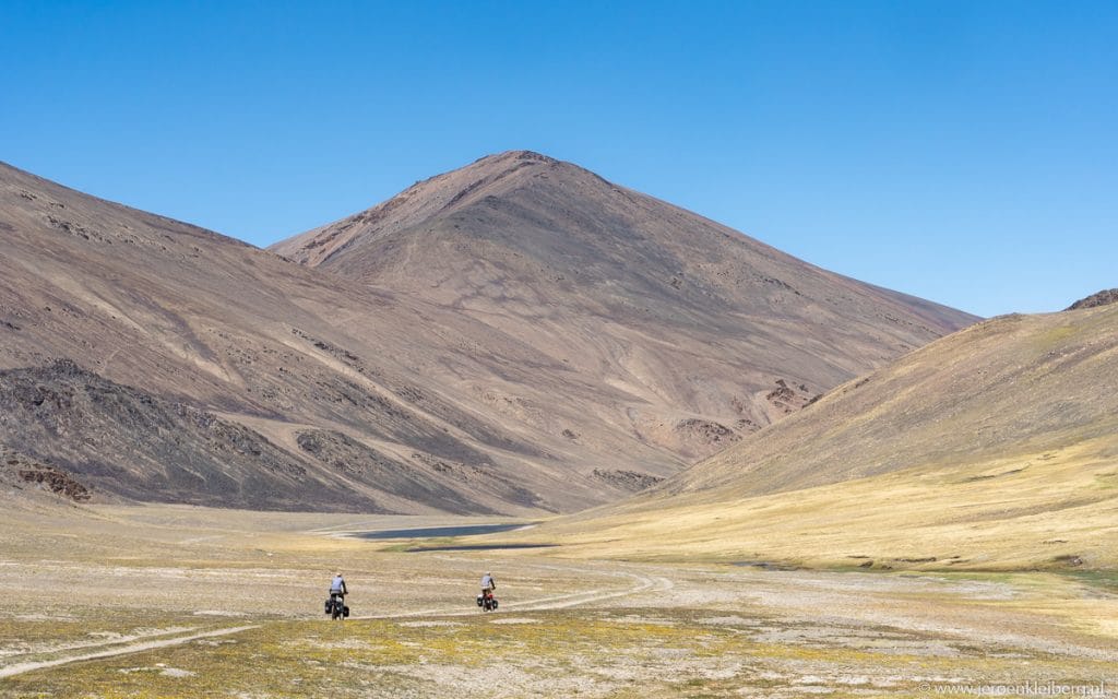 Twee fietsers met zadeltassen op een kaal hoogvlakte in de Pamir, Tadzjikistan, met bruine bergketen en blauwe lucht op de achtergrond