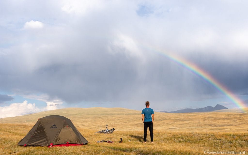 Fietser staat naast een tent en fiets op een gouden steppe in Kirgizistan en kijkt naar een grote regenboog in een dramatische lucht