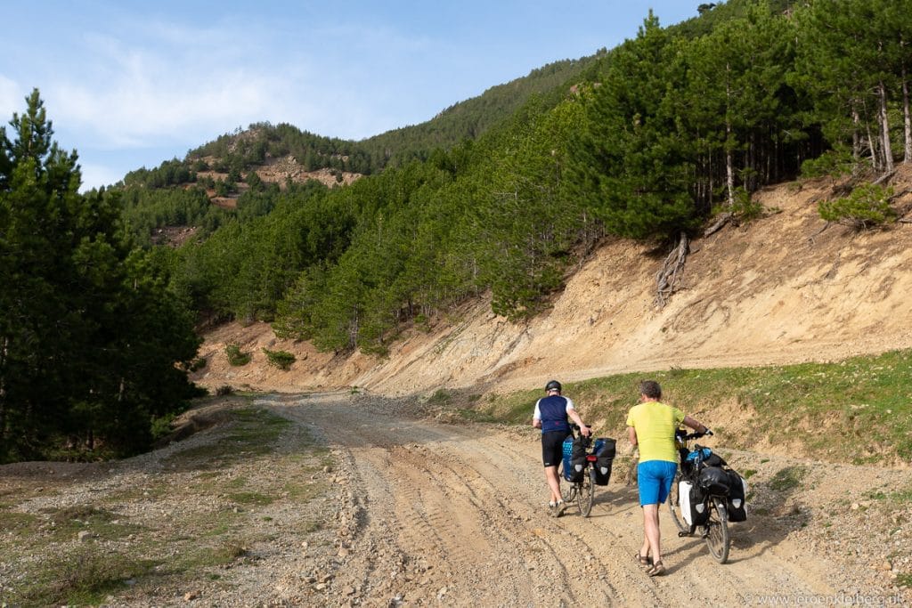 Twee fietsers met bepakking duwen hun fietsen omhoog over een steil onverhard bergpad in Albanië, met dennenbos en rotsachtige hellingen