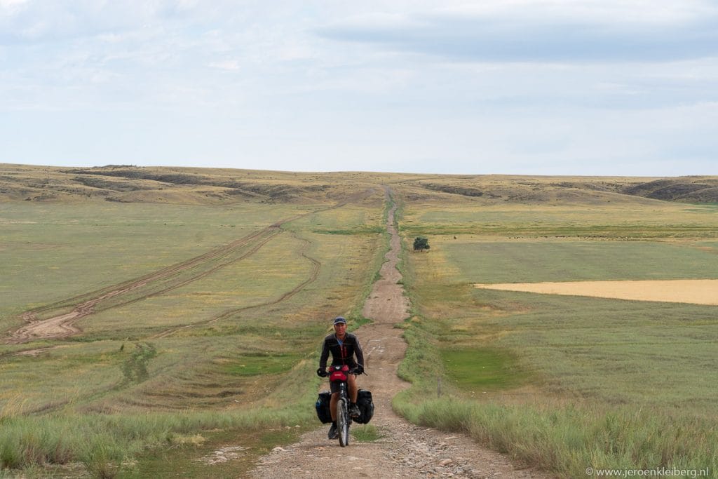 Fietser met bepakking rijdt over onverhard zandpad door de uitgestrekte Kazachstaanse steppe met vlakke graslanden en blauwe lucht