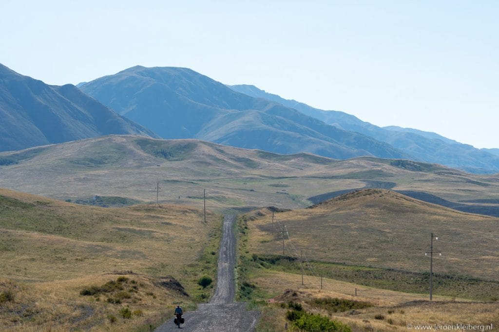 Eenzame fietser op een rechte onverharde weg door de uitgestrekte steppe van Kazachstan met een bergketen op de achtergrond