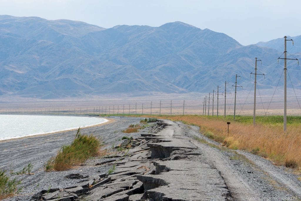 Volledig ingestort en gebarsten wegdek langs een wateroever in Kazachstan met elektriciteitspalen en bergen op de achtergrond