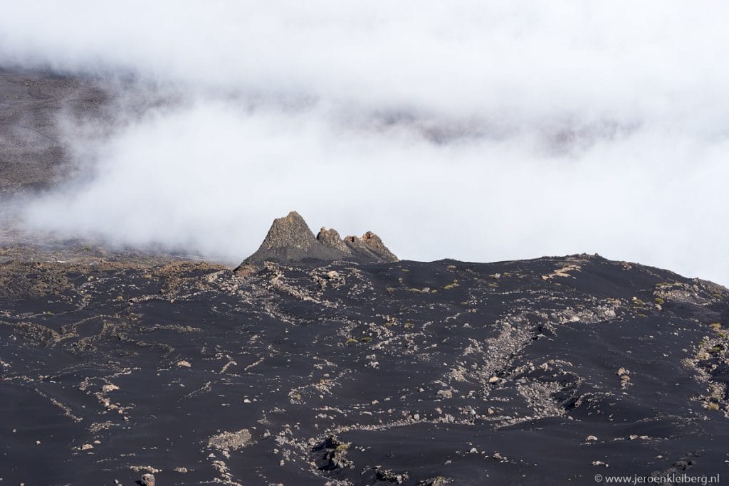 Rotsachtige pieken steken uit boven het zwarte lavalandschap van de Pico do Fogo, omhuld door lage wolken en mist op eiland Fogo in Kaapverdië