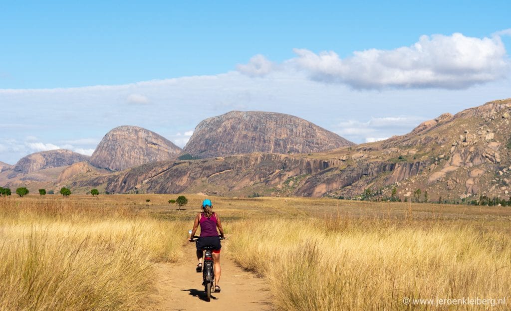 Vrouw op mountainbike rijdt op onverhard pad door graslandschap in Madagaskar met grote granieten rotsdomes op de achtergrond