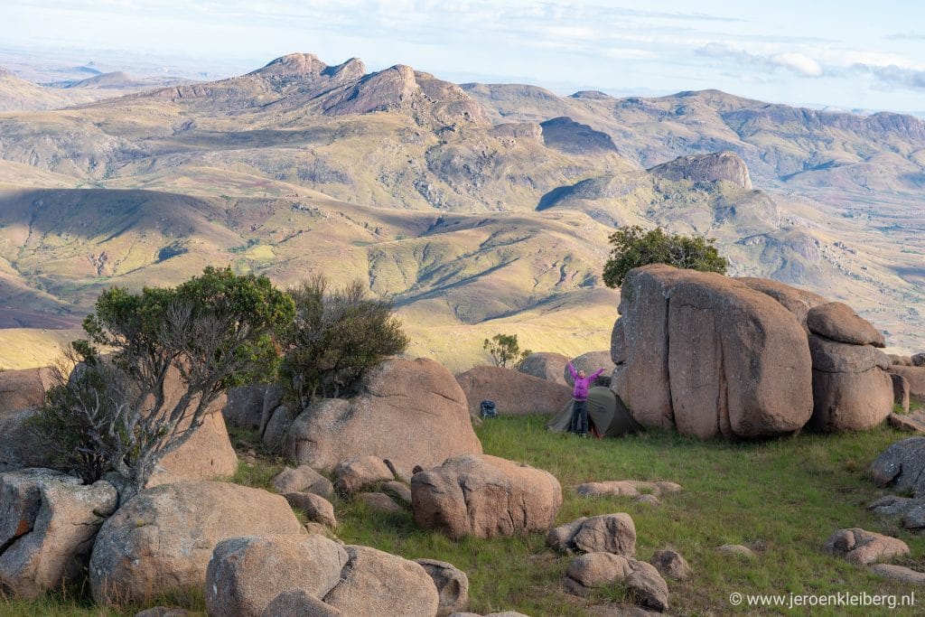 Vrouw staat met gespreide armen bij tent en granieten rotsblokken in berglandschap van Madagaskar, uitgestrekte heuvels op de achtergrond