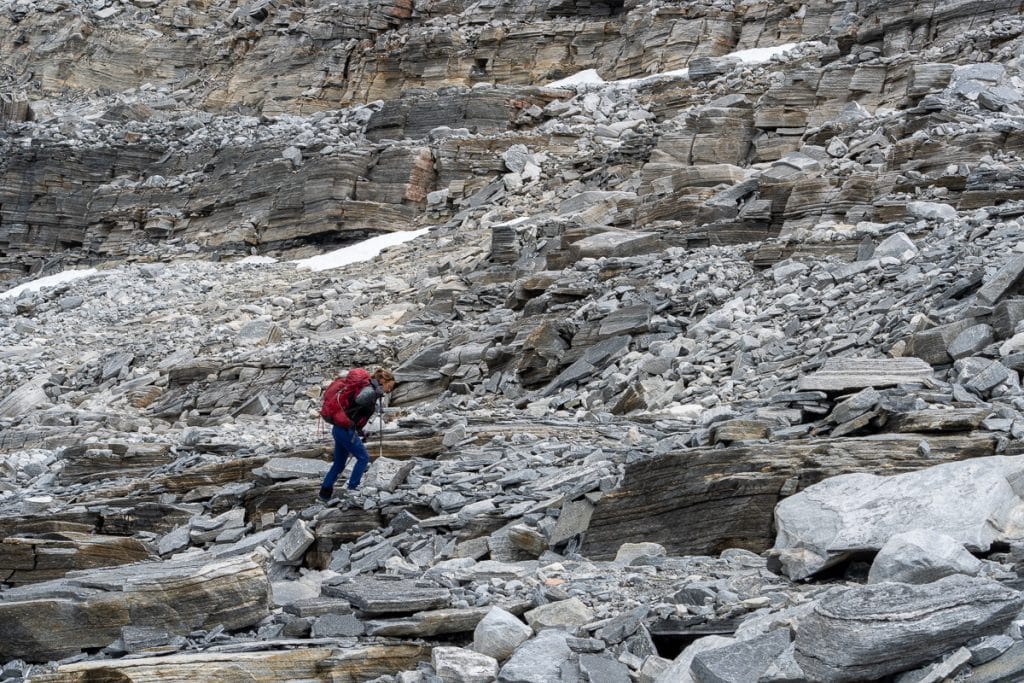 Wandelaar met rode jas en trekkingstokken klautert voorzichtig over rotsachtige helling van grijze leisteen in de Noorse fjell