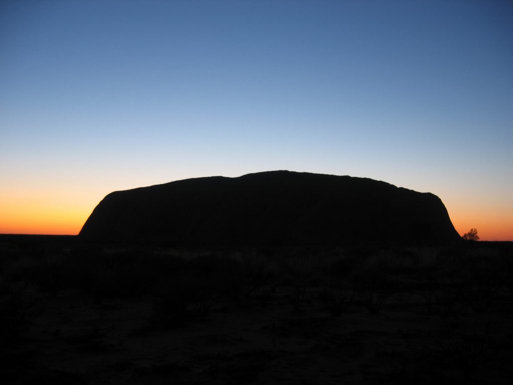 Uluru als donker silhouet bij zonsondergang tegen een oranje en blauwe hemel in het Northern Territory, Australië