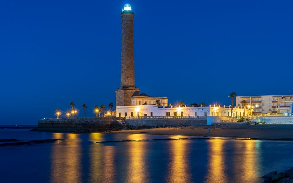 Vuurtoren van Chipiona verlicht tijdens het blauwe uur, met reflecties op het water en de Andalusische kust op de achtergrond