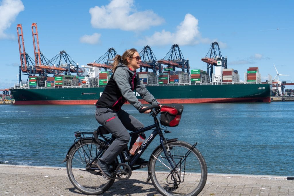 Vrouw fietst langs het havenpad in Rotterdam met een groot containerschip en rode havencranes op de achtergrond