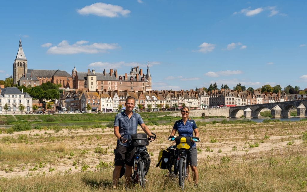 Twee fietsers met toerfietsen op de Loire-oever bij Gien, met het kasteel van Gien, de kerk en rij huizen langs de rivier op de achtergrond