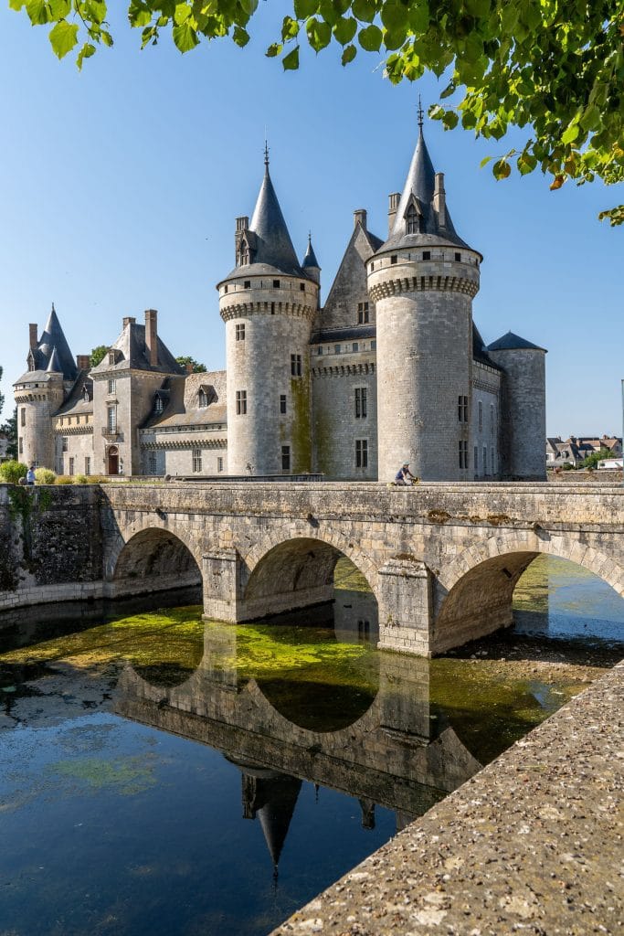 Château de Sully-sur-Loire met ronde torens en grijze spitsdaken, gedeeltelijk gekadreerd door bladeren, langs de Loire in Centre-Val de Loire