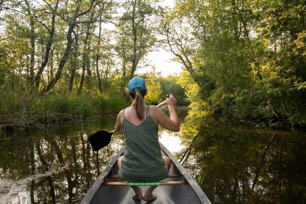 Vrouw van achteren gezien die peddelt in een kano op een smal kanaal in Giethoorn, omgeven door weelderig groen