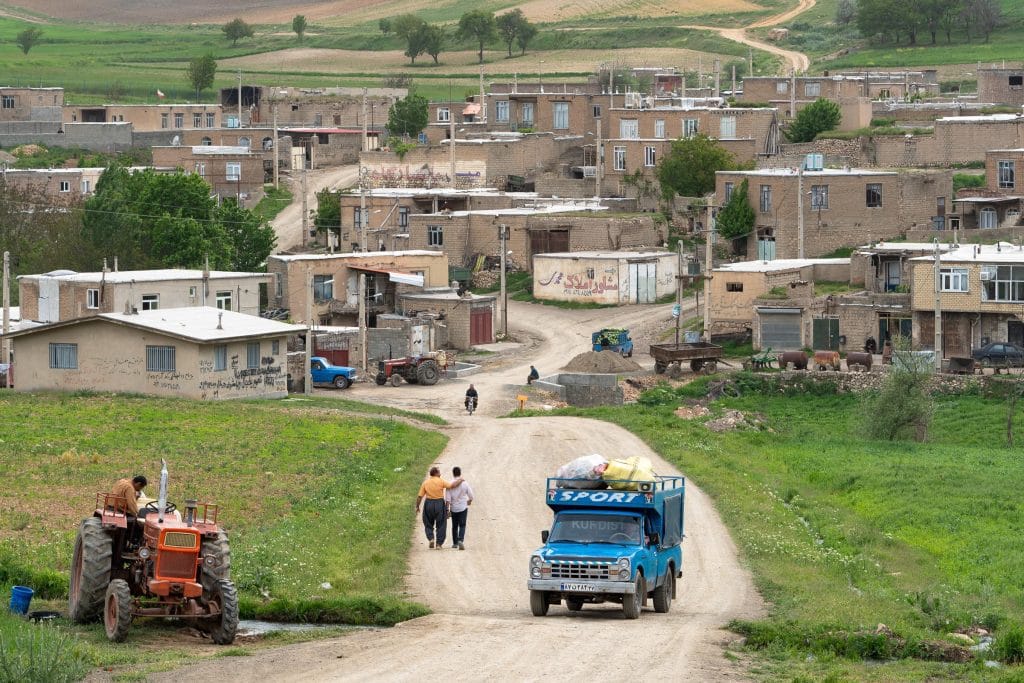 Dorpsstraat in Koerdistaans Iran met lemen huizen, een tractor en mensen op een aarden weg tussen groene heuvels