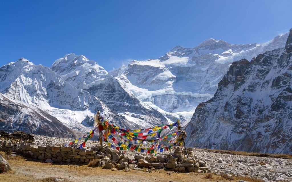 Kleurrijke gebedsvlaggen in de voorgrond, gespannen tussen steenstapels, met de adembenemende, sneeuwbedekte bergtoppen van de Himalaya op de achtergrond, onder een onberispelijk blauwe hemel, een iconisch beeld dat zowel de spirituele als de natuurlijke schoonheid van het Kanchenjunga trekpad benadrukt.