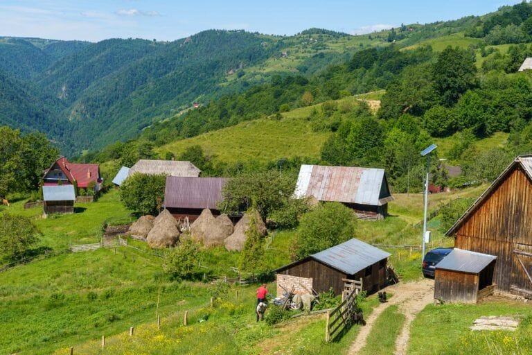 Een schilderachtig dorp in de Apuseni-bergen, Roemenië, met houten huizen, hooioppers, groene heuvels en een boer in een rode trui die buiten werkt.