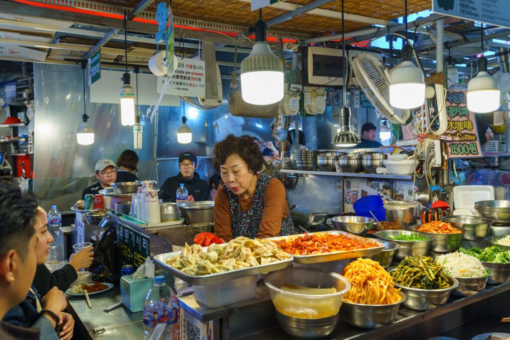 Een drukke kraam op een Koreaanse markt waar een vrouw gerechten bereidt, omringd door schalen vol kleurrijke ingrediënten en klanten die genieten van een snelle maaltijd.