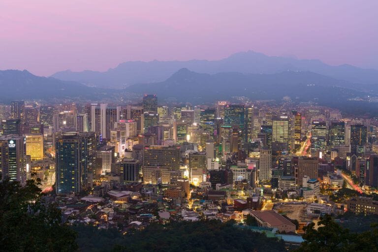 Panoramisch uitzicht op de skyline van Seoul bij zonsondergang, met verlichte gebouwen en bergketens aan de horizon.