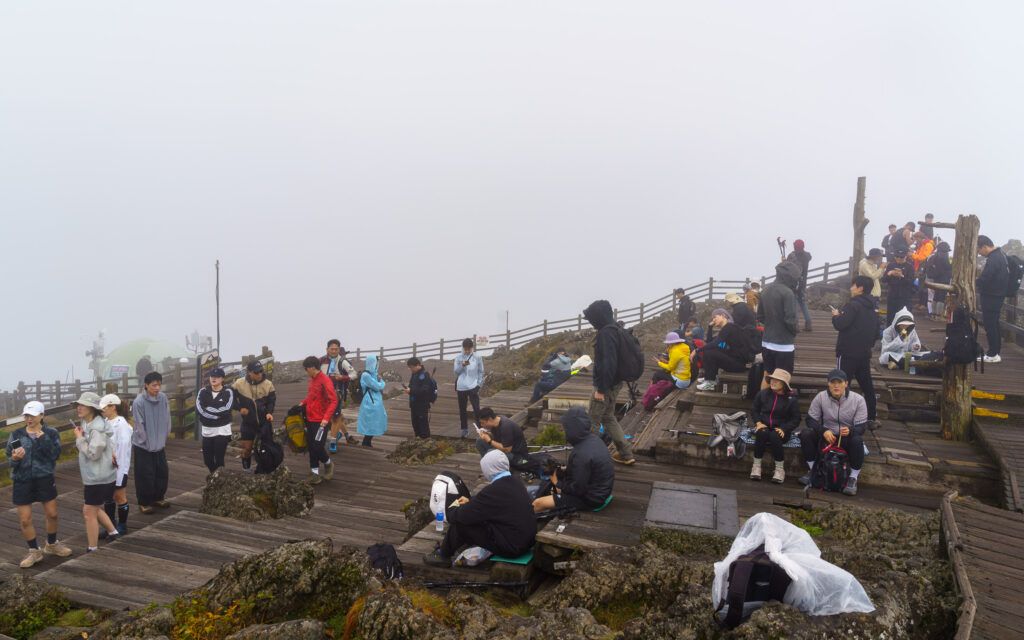 Groep wandelaars op een mistige bergtop, zittend en staand op houten platforms, gehuld in high-tech outdoor kleding.