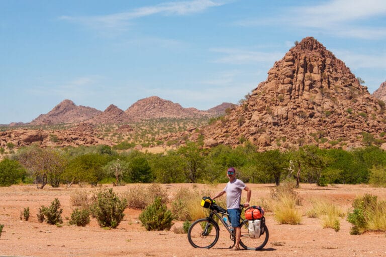 Fietser staat met beladen toerfiets voor een piramidaal granieten bergmassief in droog Namibisch landschap