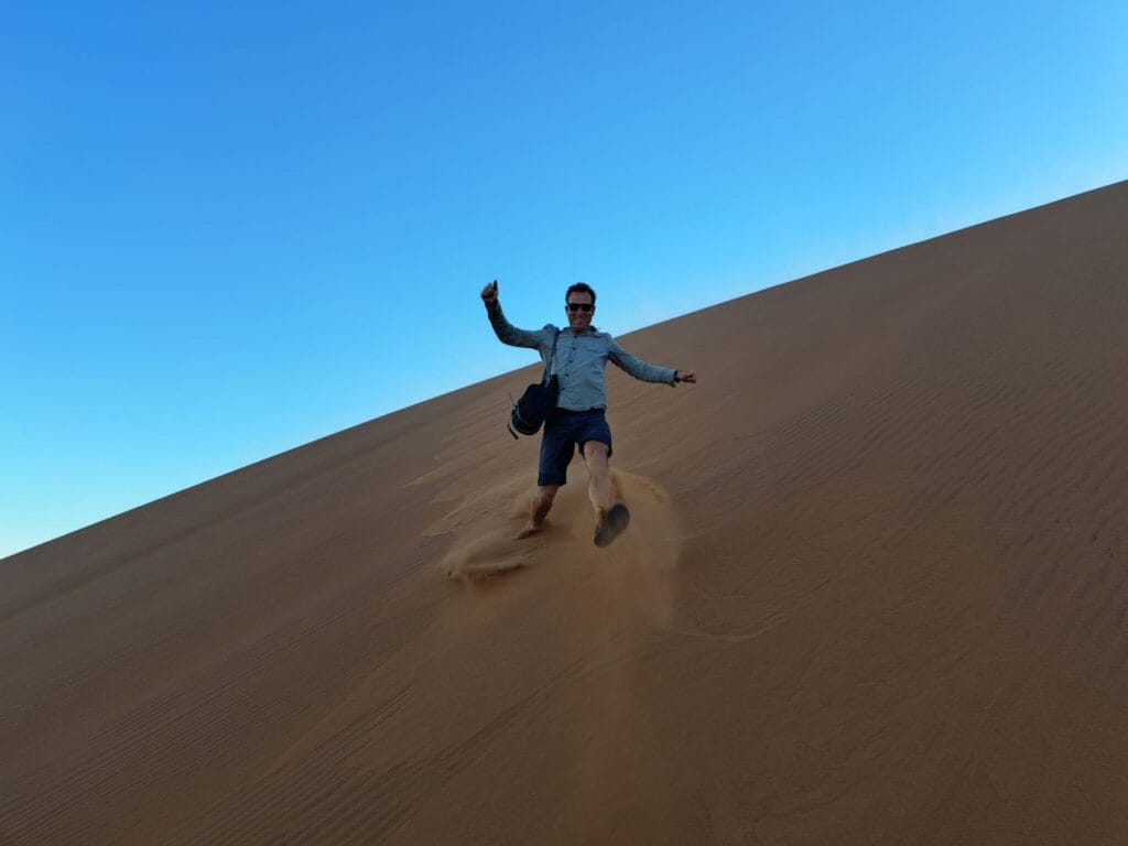 Man met zonnebril en camera loopt lachend en met armen gespreid naar beneden langs een grote oranje zandduin in Namibia onder strakblauwe lucht