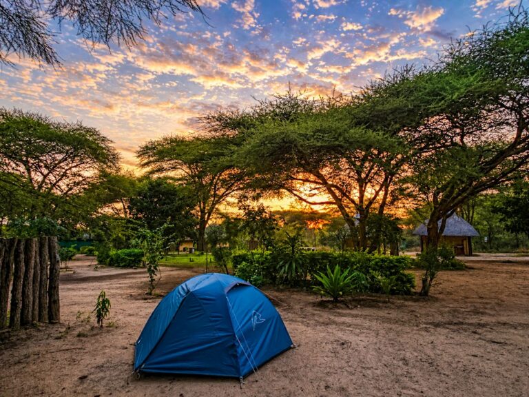 Blauwe koepeltent op een zandige campsite tussen acaciabomen bij zonsondergang, met een traditionele rondavel op de achtergrond