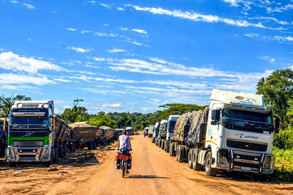 Lange rij grote vrachtwagens met cargo langs een onverharde weg in de Zambiaanse bush, met een fietser ertussen