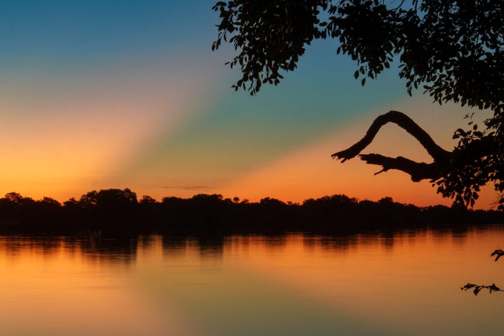 Vlammend oranje zonsondergang boven Lake Kariba in Zimbabwe met boomsilhouet op de voorgrond en rustige weerspiegeling in het water