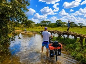Fietser duwt een bepakte toerfiets door een ondiepe rivier bij een primitieve houten brug in zuidelijk Afrika, een ander persoon loopt voorop