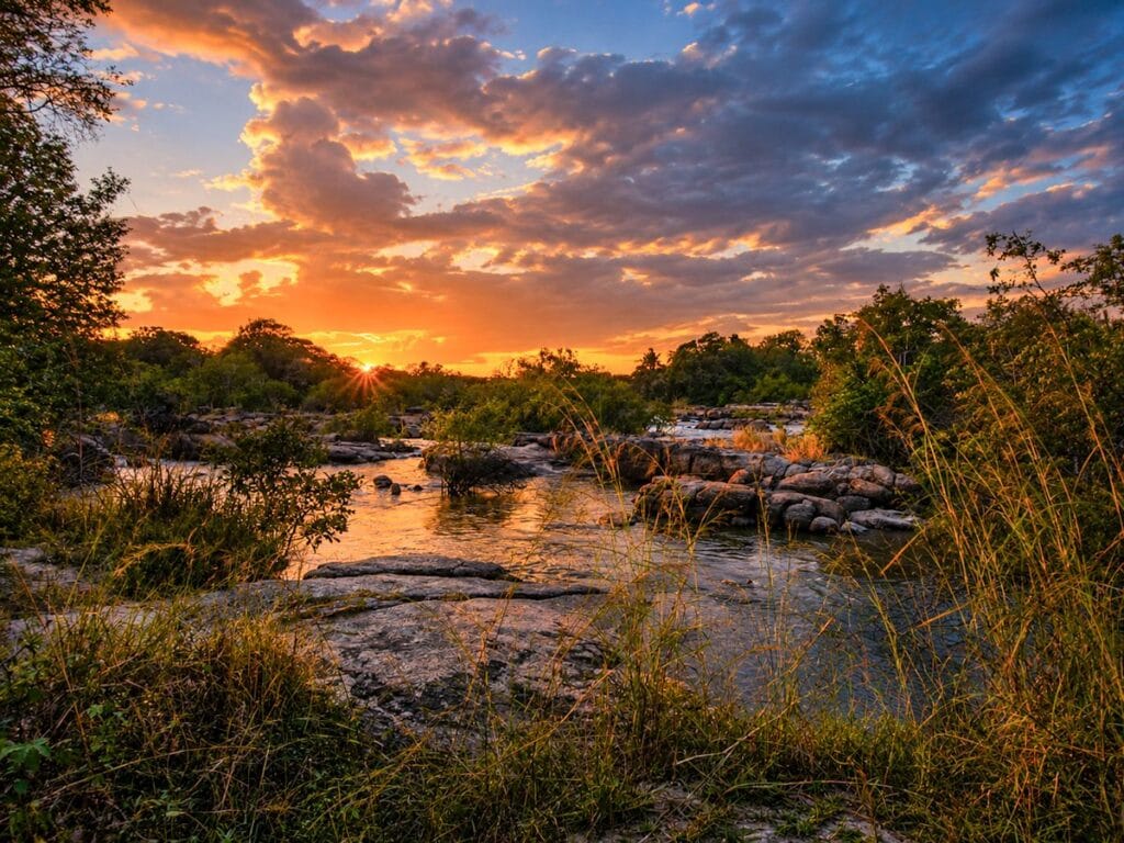 Vurige oranje zonsondergang boven een ondiepe rotsige rivier met rietgras en struikgewas in Zimbabwe