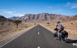 Kaarsrechte asfaltstrip door een kale vallei met verspreide acacias en een beladen reisfietser
