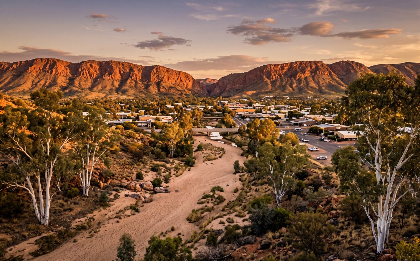 Alice Springs in een dal tussen de rode MacDonnell Ranges met de droge zanderige Todd River en witte gum-bomen in gouden avondlicht