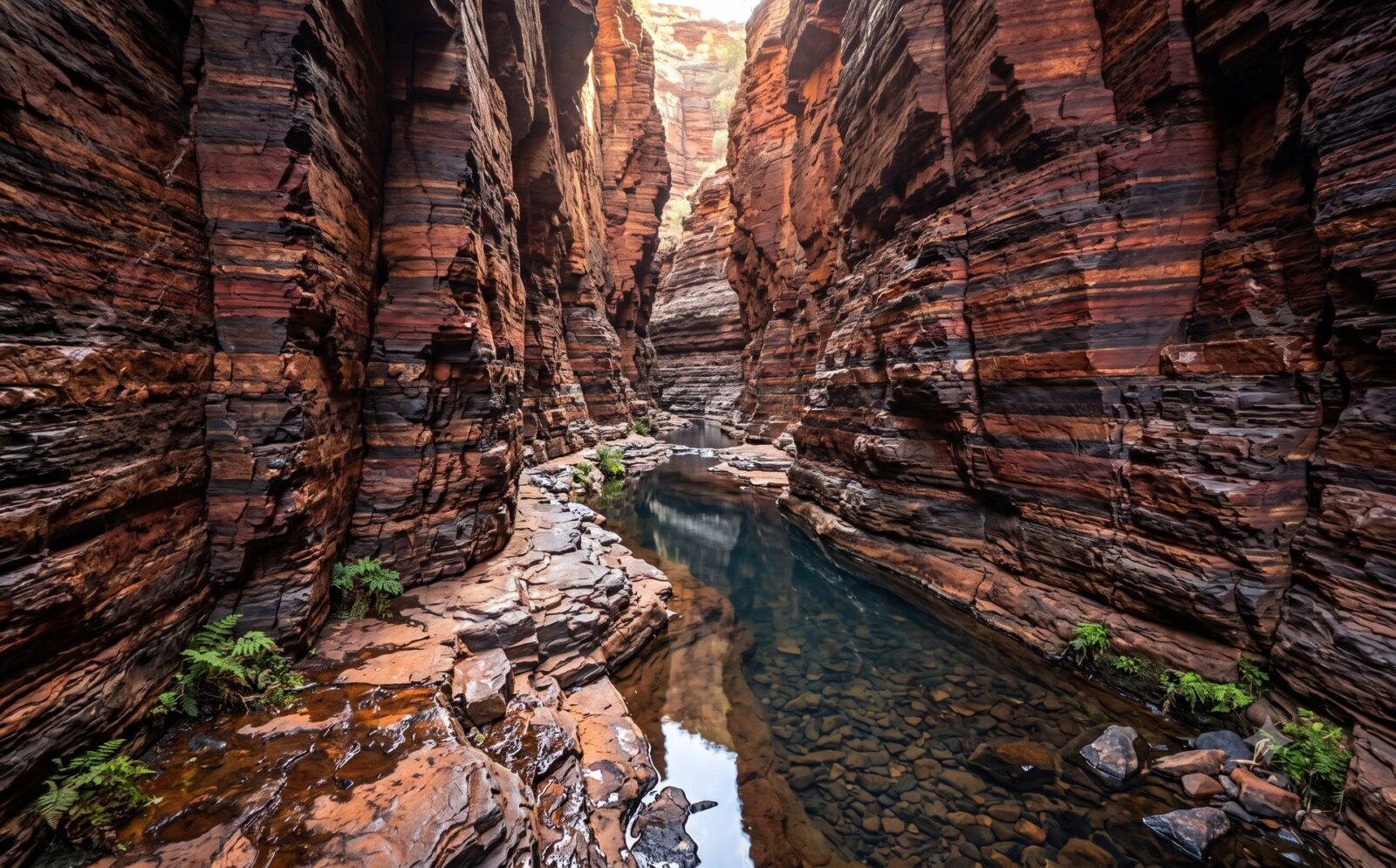 Diepe gorge in Karijini met loodrechte roodzwart gestreepte gesteentewanden en een smalle waterpoel op de bodem