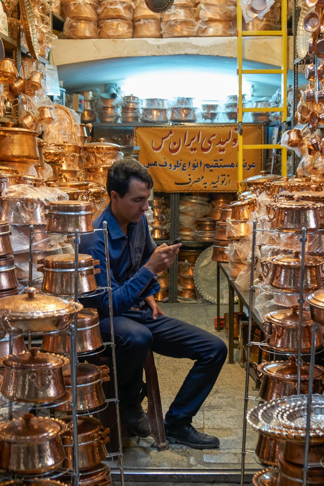 Man kijkt op zijn telefoon tussen stapels koperwerk in Yazd