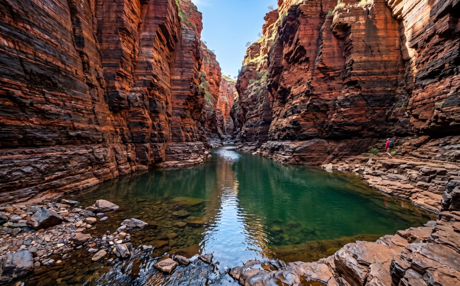 Smalle diepe gorge in Karijini met loodrechte roodzwart gestreepte wanden en een heldere waterpoel op de bodem in gefilterd zonlicht