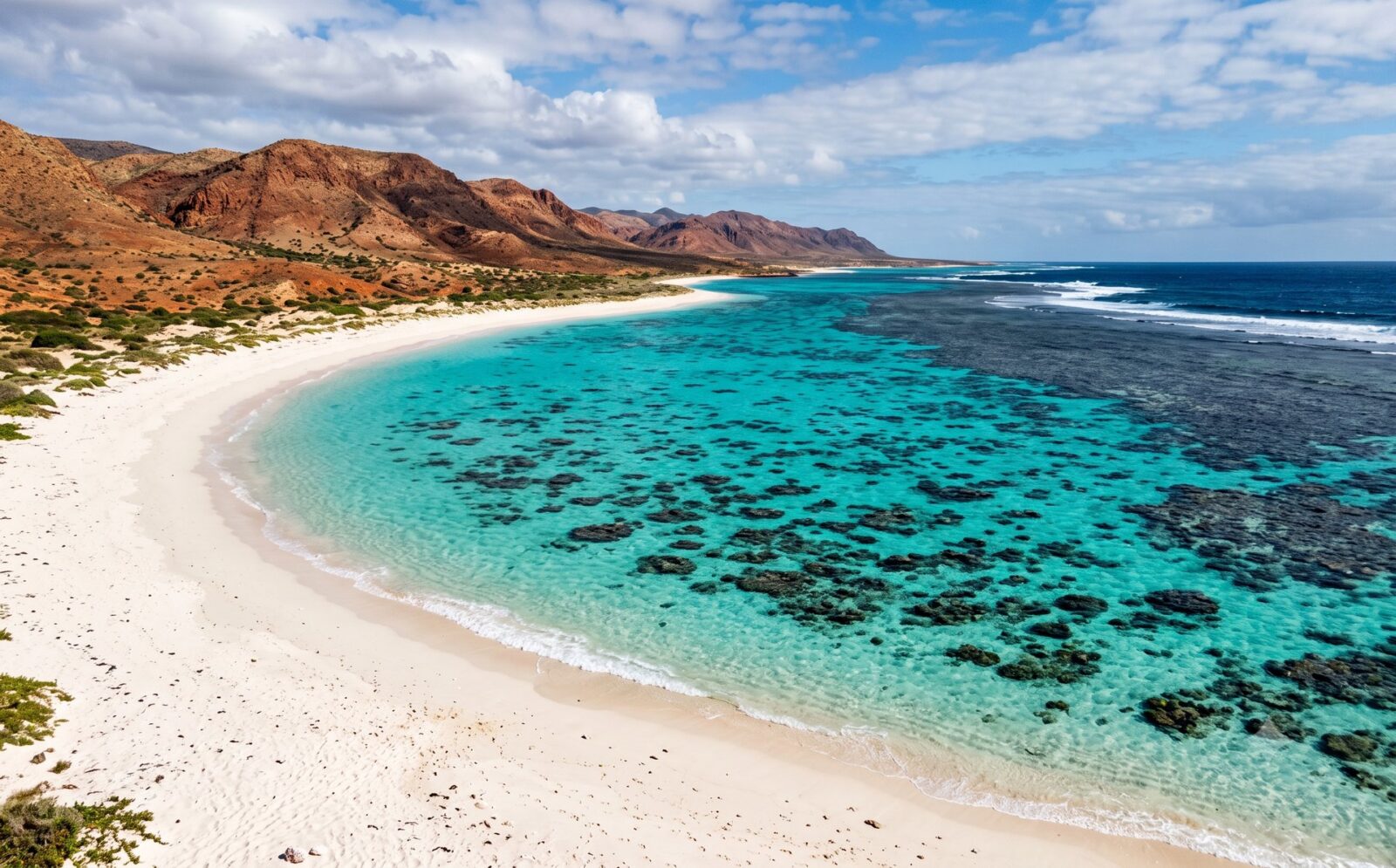Kust bij Ningaloo waar een wit strand overgaat in een turquoise lagune met zichtbaar koraal en droge rode heuvels van het Cape Range erachter