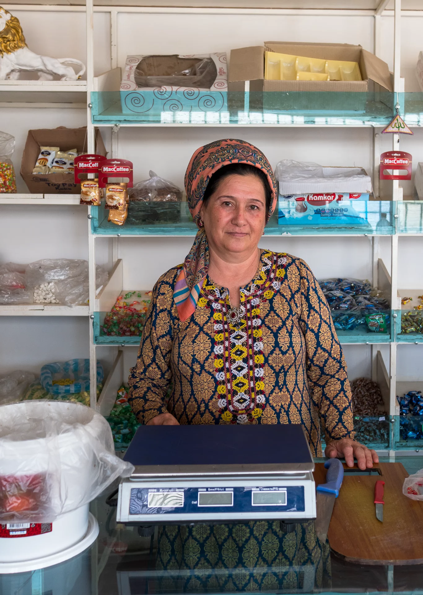 Vrouw in donkerblauwe geborduurde jurk achter de weegschaal in een winkel in Sarakhs